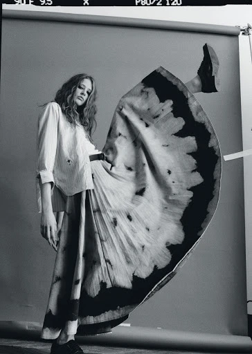 Fashion shot of a woman in a long skirt and collared shirt posing with one leg up in a studio with a gray background.