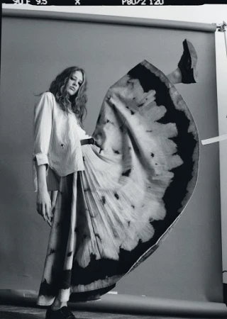 Fashion shot of a woman in a long skirt and collared shirt posing with one leg up in a studio with a gray background.
