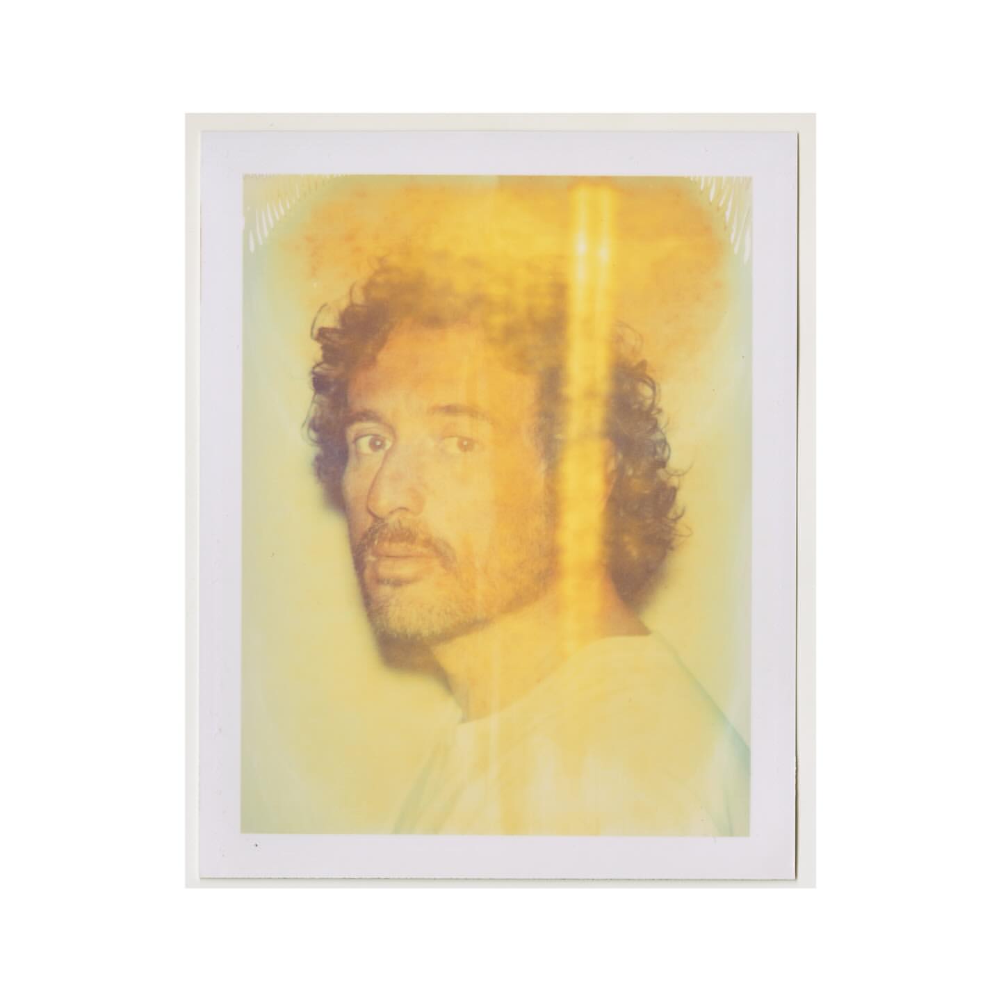 Portrait of an adult male with curly hair and a beard, wearing a white shirt, in a photograph with an abstract yellow and white background.