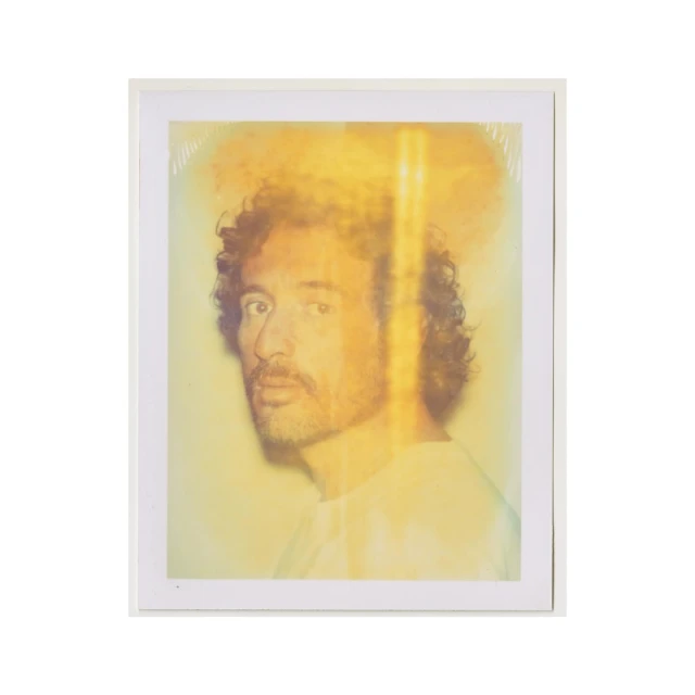 Portrait of an adult male with curly hair and a beard, wearing a white shirt, in a photograph with an abstract yellow and white background.