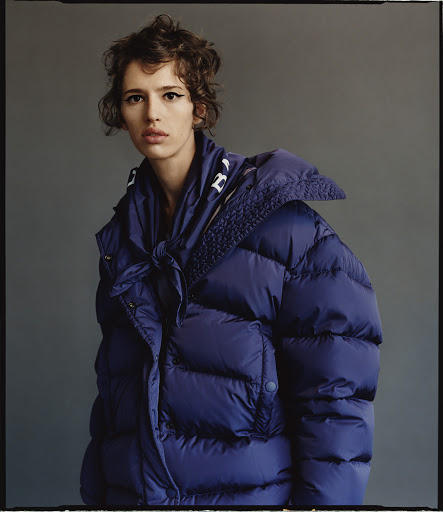 A woman with short curly brown hair poses in a studio wearing a large blue puffer jacket with a white logo on the collar.