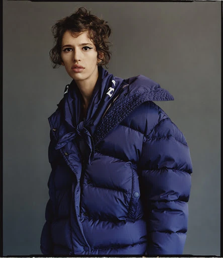 A woman with short curly brown hair poses in a studio wearing a large blue puffer jacket with a white logo on the collar.