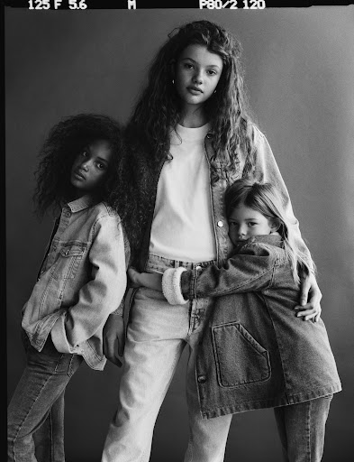 Three young girls pose together in denim outfits in a monochrome studio shot. They wear jeans and jackets, their hair styled naturally.