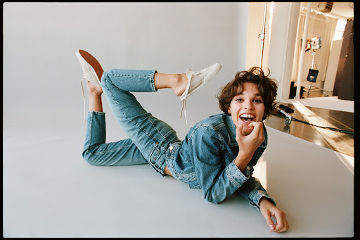 Adult female lying on white surface wearing jeans, denim jacket, and sneakers. The model is smiling and looking at the camera.