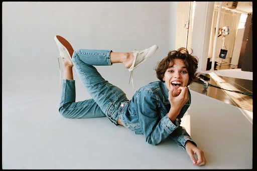 Adult female lying on white surface wearing jeans, denim jacket, and sneakers. The model is smiling and looking at the camera.
