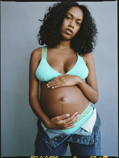 Pregnant woman with dark curly hair posing indoors wearing a teal top, and denim jeans. Fashion shot of a mother-to-be.