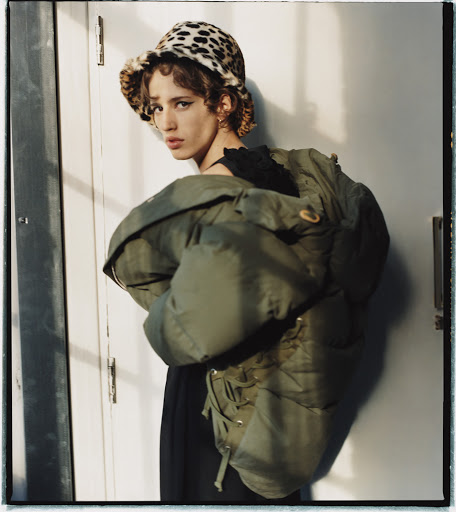 Stylish young adult woman poses indoors wearing an oversized green puffer jacket and a leopard print bucket hat, eye-level angle.