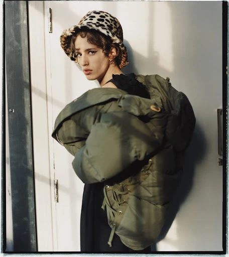Stylish young adult woman poses indoors wearing an oversized green puffer jacket and a leopard print bucket hat, eye-level angle.