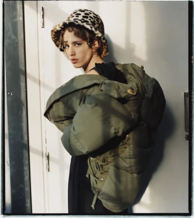 Stylish young adult woman poses indoors wearing an oversized green puffer jacket and a leopard print bucket hat, eye-level angle.