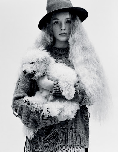 Young woman with long blonde hair and a hat holding a white poodle. She is wearing a distressed sweater in a black and white studio shot.
