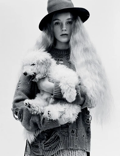 Young woman with long blonde hair and a hat holding a white poodle. She is wearing a distressed sweater in a black and white studio shot.