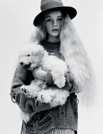 Young woman with long blonde hair and a hat holding a white poodle. She is wearing a distressed sweater in a black and white studio shot.