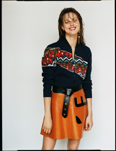 Smiling woman with a wet hair look poses in a studio wearing a printed zip-up jacket, a tan leather skirt, and dangling earrings.
