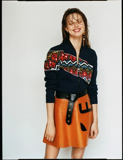 Smiling woman with a wet hair look poses in a studio wearing a printed zip-up jacket, a tan leather skirt, and dangling earrings.