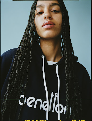 Close-up studio shot of a young woman with braided hair and a septum piercing, wearing a dark hoodie against a light blue backdrop.