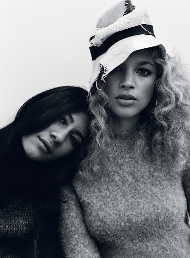 Two women posing closely together in monochrome, one wearing a textured sweater and a light-coloured brimmed hat.