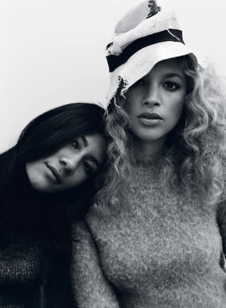 Two women posing closely together in monochrome, one wearing a textured sweater and a light-coloured brimmed hat.