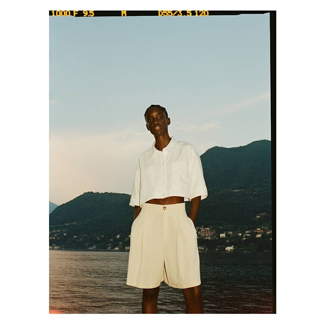 Outdoor fashion shot of a Black woman in white blouse and beige shorts, standing near a lake with mountains in the background.