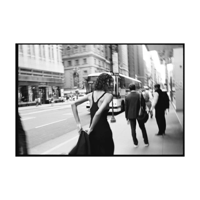 Black-and-white street style: a woman in a black dress and jacket walks along a city sidewalk with office buildings and a bus in the background.