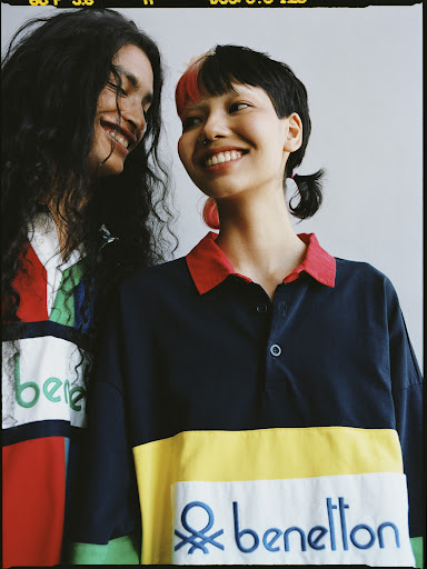 Two happy adult female models smile, wearing Benetton rugby shirts in a close-up studio shot against a neutral background. Fashion editorial.