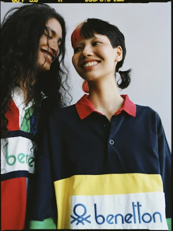 Two happy adult female models smile, wearing Benetton rugby shirts in a close-up studio shot against a neutral background. Fashion editorial.