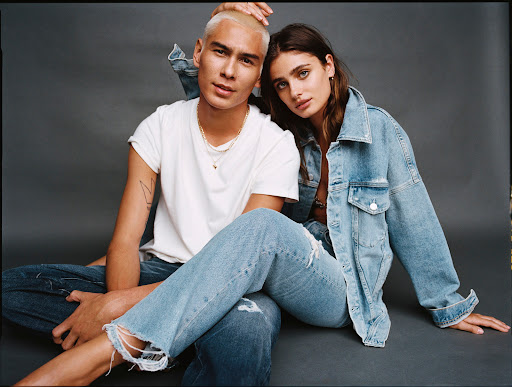 A couple models denim fashion, posing seated on a gray background. The man with buzzcut hair wears a white t-shirt and jeans; the woman a denim jacket.
