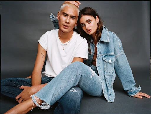 A couple models denim fashion, posing seated on a gray background. The man with buzzcut hair wears a white t-shirt and jeans; the woman a denim jacket.