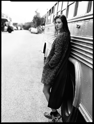 Black and white portrait of a young woman leaning against a bus wearing a speckled coat and dark skirt in an urban environment.