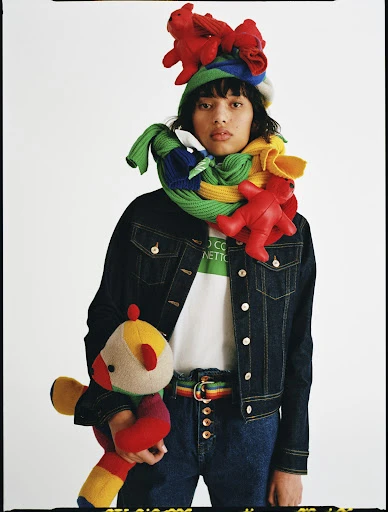 Person wearing a multicolored scarf, hat with stuffed animals, denim jacket and jeans, holding a stuffed animal, white background.