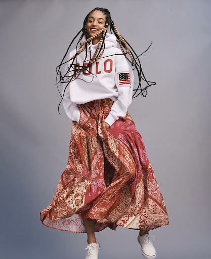 Young woman with braided hair, wearing a Polo hoodie and patterned skirt, jumps in a studio shot on a grey background.
