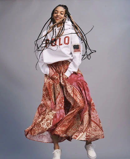 Young woman with braided hair, wearing a Polo hoodie and patterned skirt, jumps in a studio shot on a grey background.