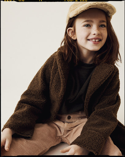 Young girl poses wearing a brown teddy bear coat, tan corduroy pants, and a newsboy cap, set against a neutral backdrop in a portrait.