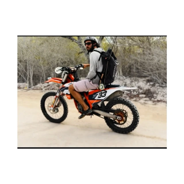 Man on orange and white dirt bike wearing a light-colored shirt, backpack, sunglasses, and shorts riding on a dirt path.