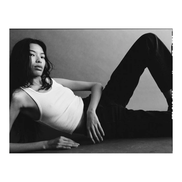A young Asian woman with long, dark hair is lying down wearing a white tank top and dark pants, striking a casual pose.