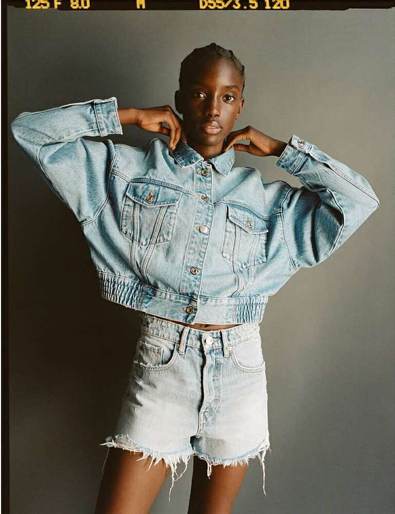 Woman in light blue denim jacket and matching shorts poses against a gray background. Fashion-forward style with neutral backdrop.