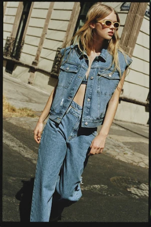 Blonde woman wearing a denim vest and matching jeans while walking on an urban street.