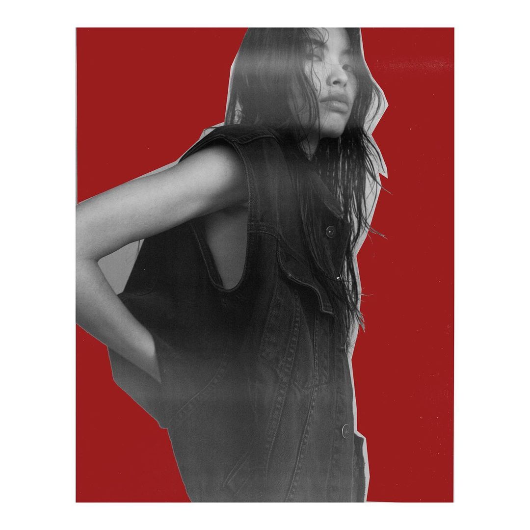 Monochrome fashion portrait of a young adult woman with long dark hair, wearing a denim vest against a red background.