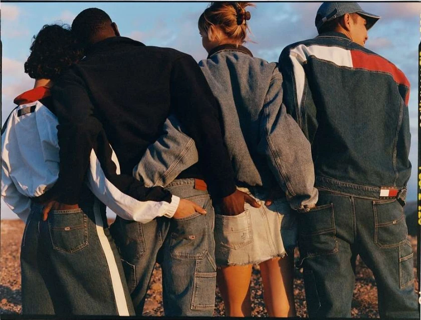Four people stand in an outdoor setting, backs facing the camera, wearing denim jackets and jeans from Tommy Hilfiger fashion brand.