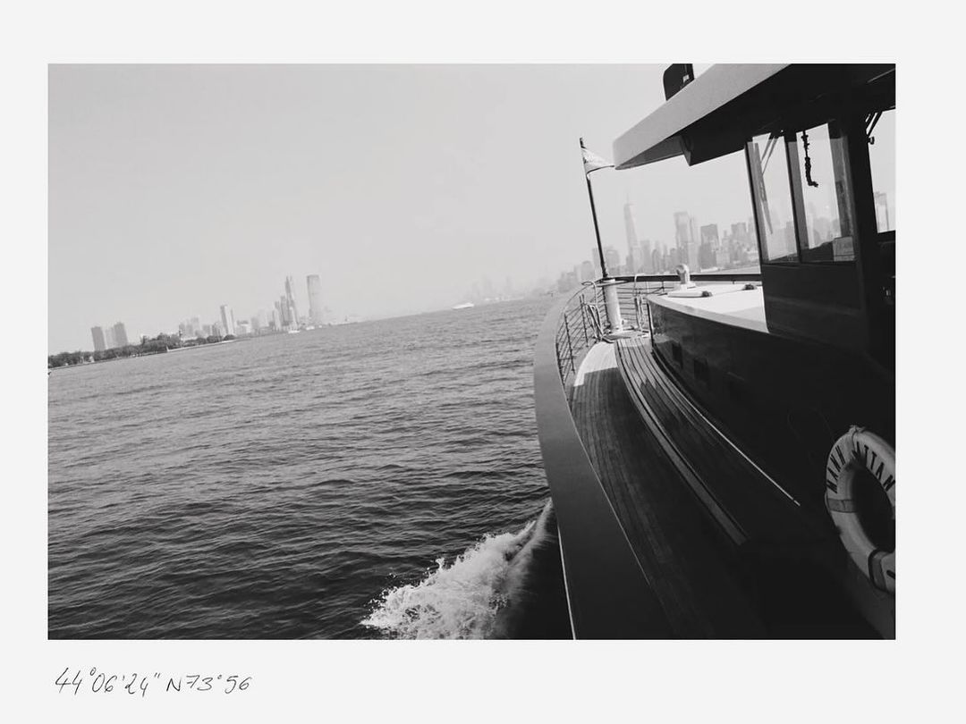 Monochrome view of a yacht sailing on the water, featuring a city skyline in the background, captured in an eye-level medium shot.