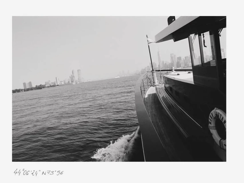 Monochrome view of a yacht sailing on the water, featuring a city skyline in the background, captured in an eye-level medium shot.