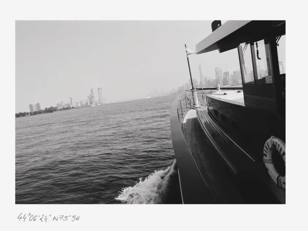 Monochrome view of a yacht sailing on the water, featuring a city skyline in the background, captured in an eye-level medium shot.