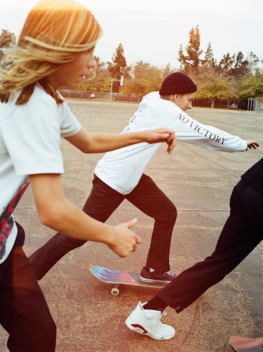 Three skateboarders ride across a concrete surface outdoors, with light bouncing off their white t-shirts, dark pants, and footwear.