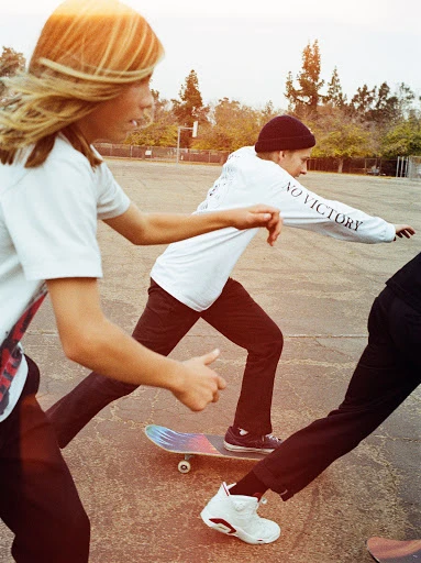 Three skateboarders ride across a concrete surface outdoors, with light bouncing off their white t-shirts, dark pants, and footwear.