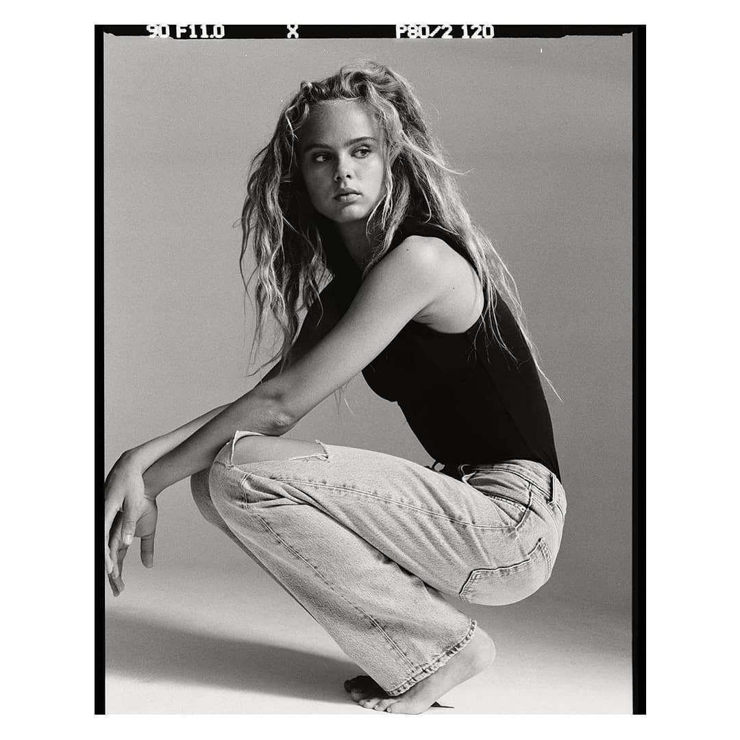 Monochrome portrait of a young woman with long tousled hair crouching in a black tank top and light ripped jeans against a seamless backdrop
