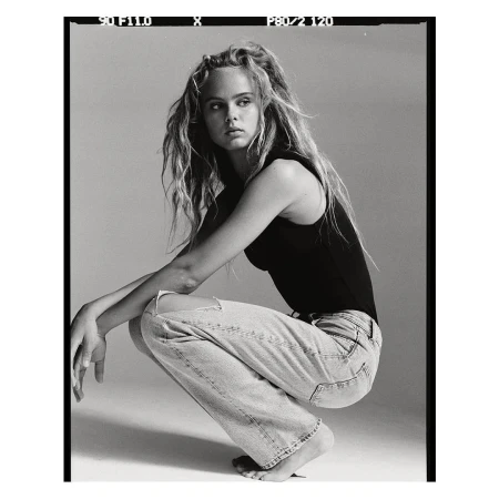 Monochrome portrait of a young woman with long tousled hair crouching in a black tank top and light ripped jeans against a seamless backdrop