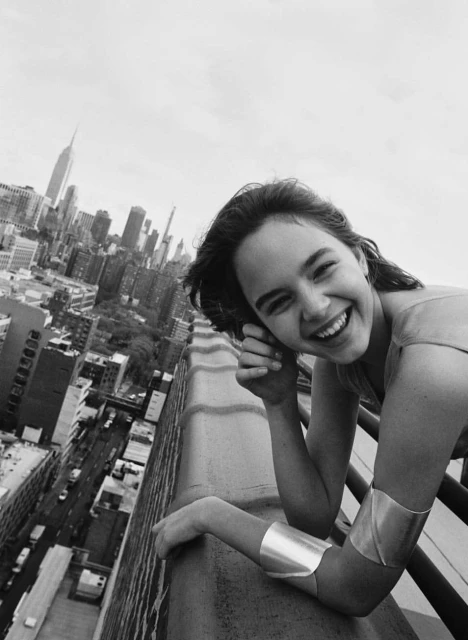 Smiling woman poses on a rooftop in an urban setting with a cityscape in the background. Black and white photography, medium shot.