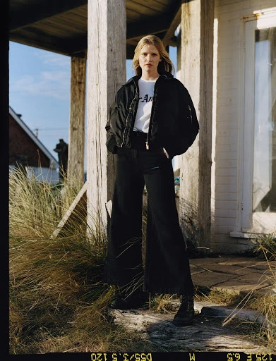 Adult woman wearing a black bomber jacket and dark, wide-leg pants poses in front of a weathered building and grassy landscape outdoors.