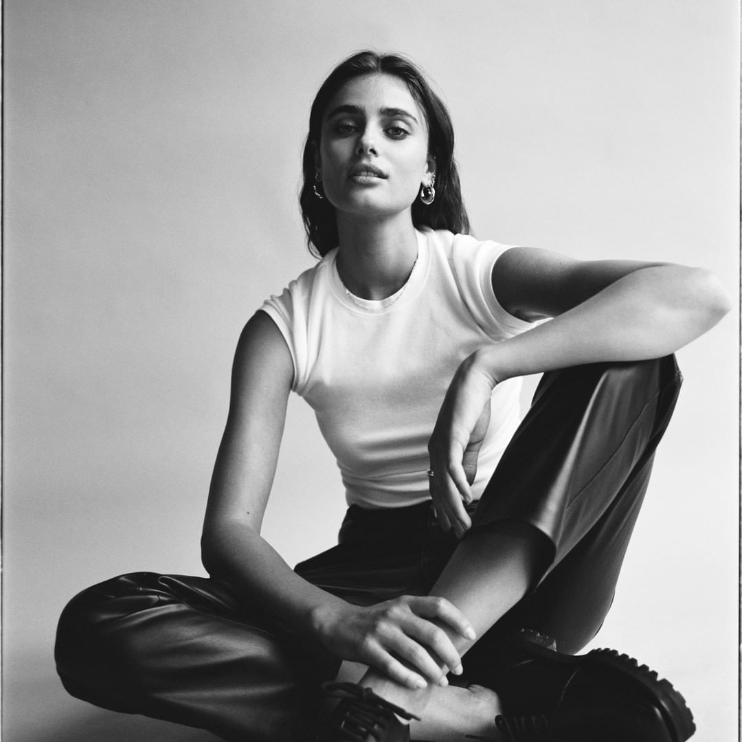 Studio portrait of an adult female with dark hair wearing a white t-shirt and dark pants, sitting with her legs crossed.