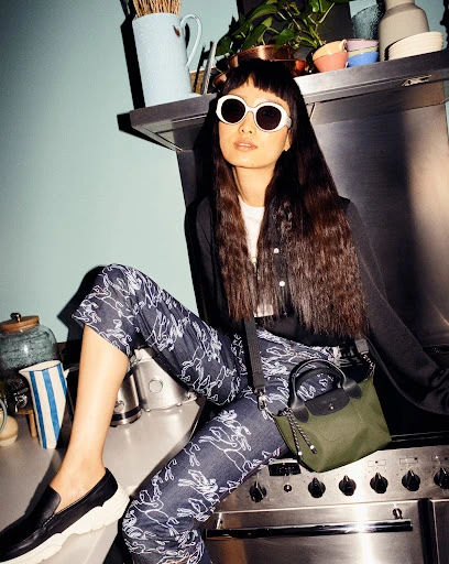 Woman with bangs and sunglasses poses on a silver stove in cropped floral pants, a black jacket, and black loafers with a green purse.