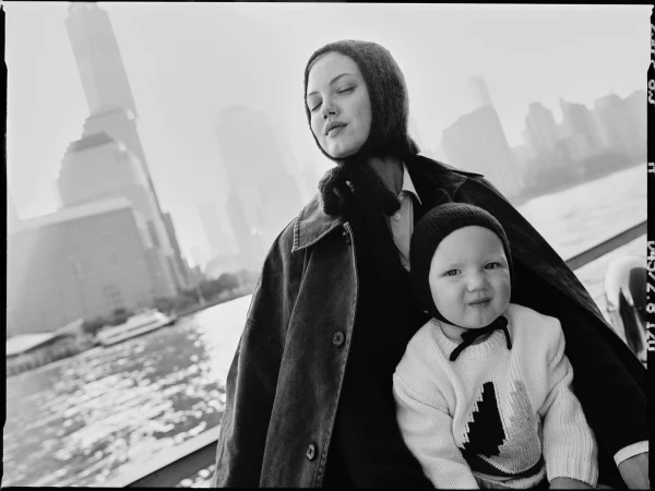 Black and white portrait of a woman in a hooded garment holding a baby with buildings and water in the background.